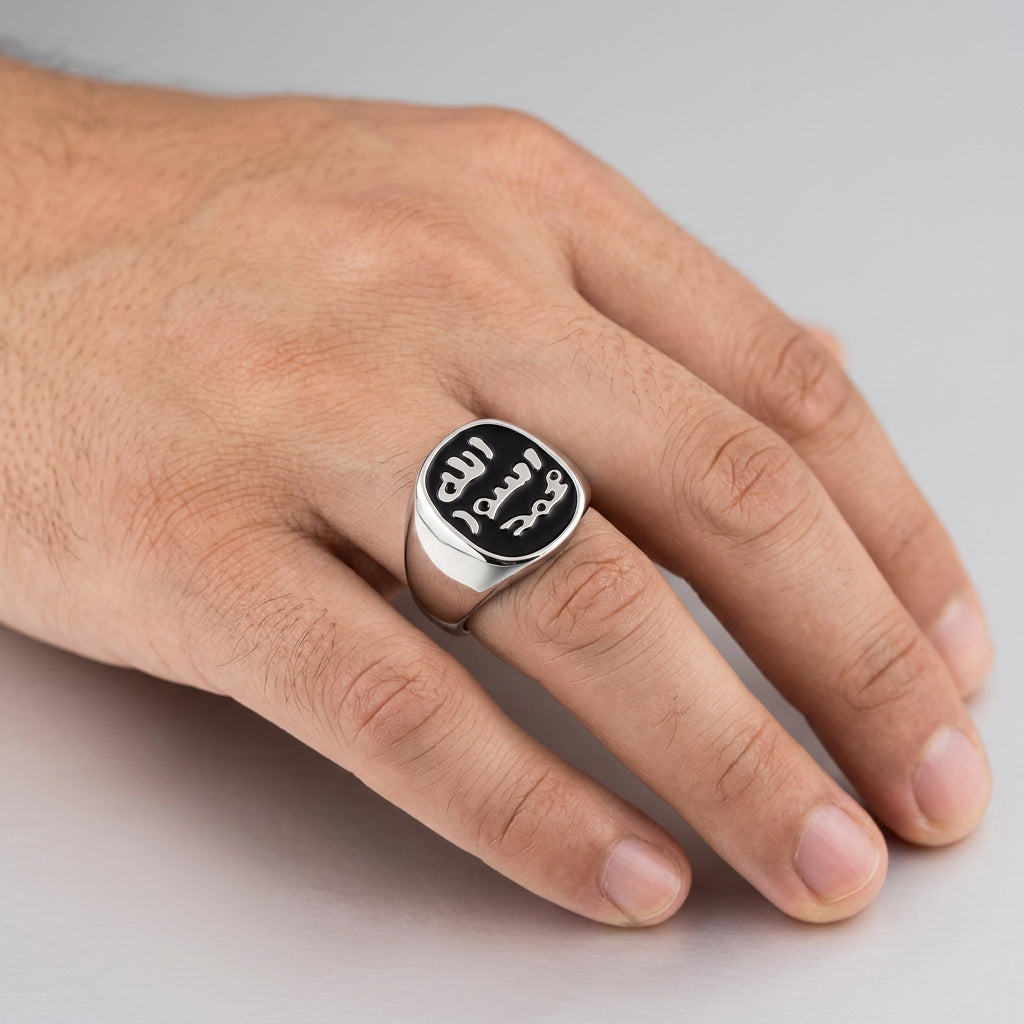 A silver-toned signet ring with Arabic calligraphy engraved on a dark oval face, shown worn on a person's finger against a dark gray background.