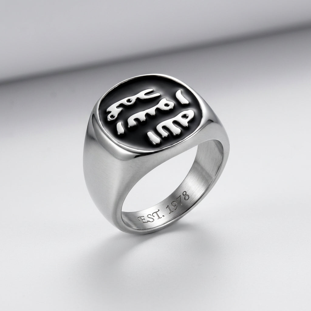 A silver signet ring with a polished oval face featuring a black inlay and stylized white cutout lettering, shown on a plain white background.