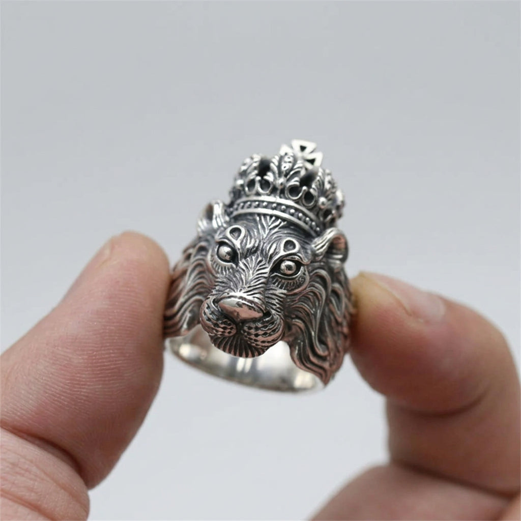 A silver lion head ring with a crown, held between two fingers against a blurred wooden background.