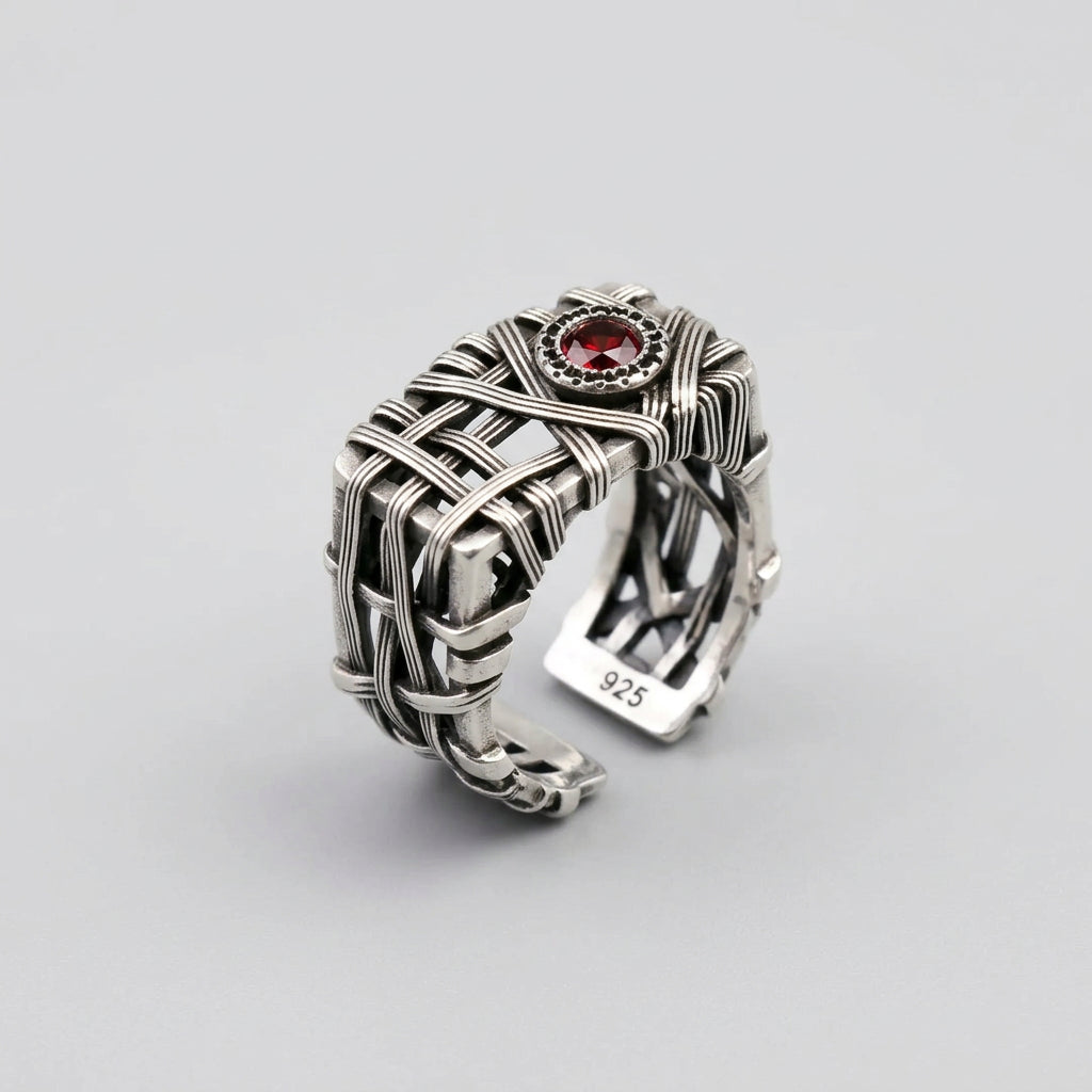 A silver-toned wrap-style ring with a small red gemstone accent, photographed on a dark textured surface.