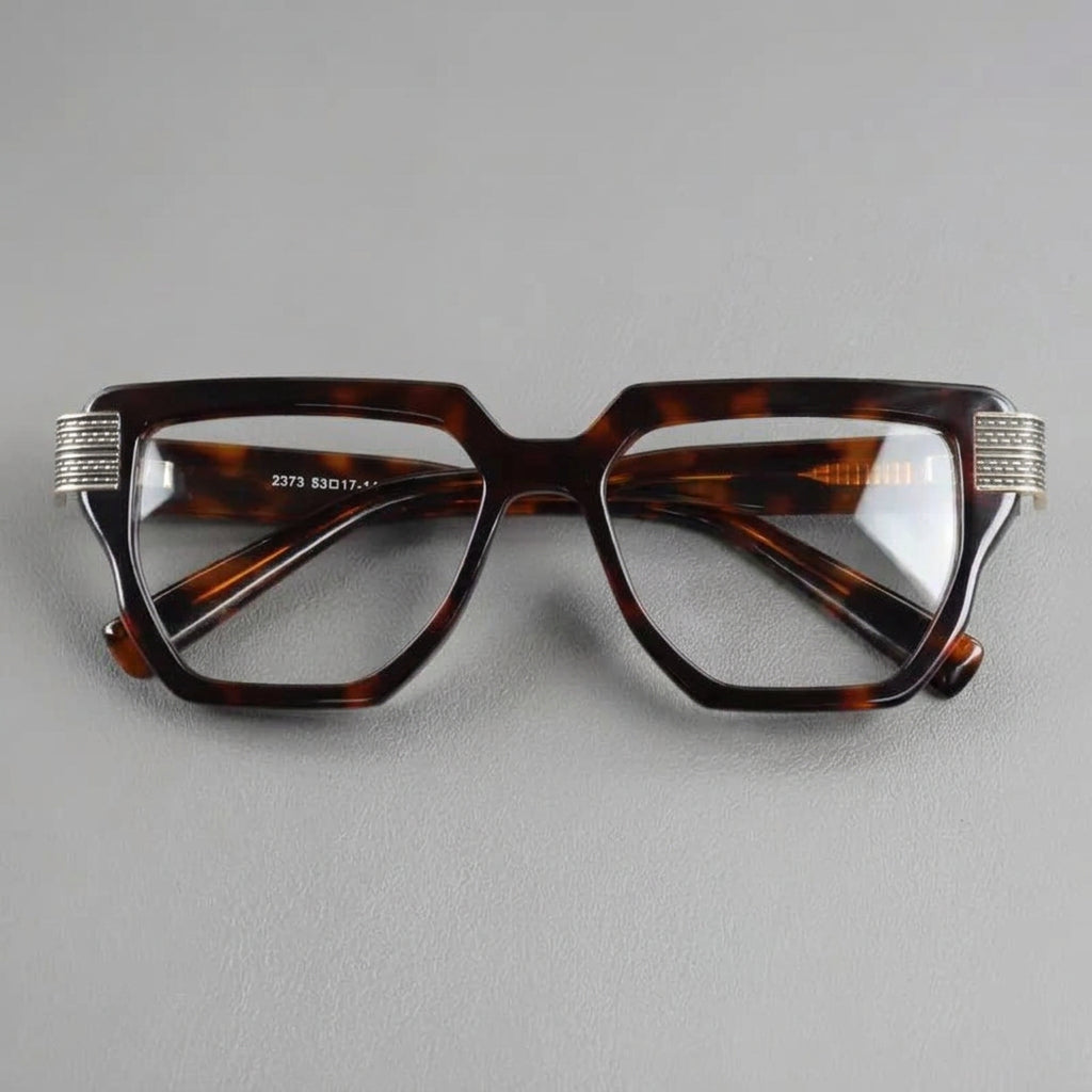 A pair of geometric square eyeglasses with a dark brown and black tortoiseshell acetate frame, featuring silver rhinestone-embellished metal tips on the temples, photographed on a textured gray background.